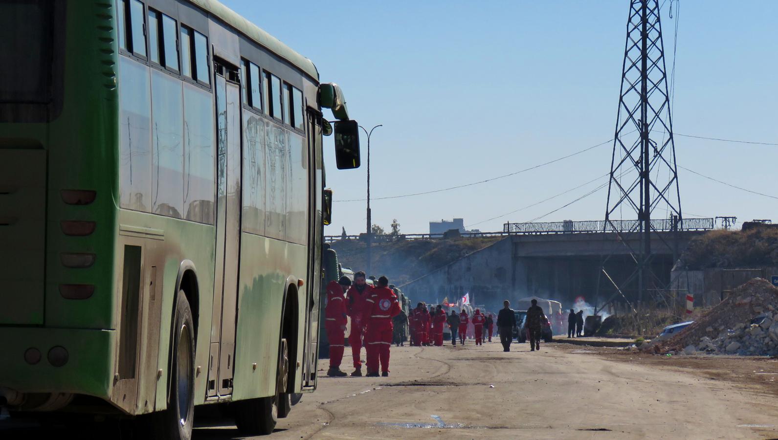 Un grupo de miembros de la Media Luna Roja espera junto a los autobuses preparados para evacuar de Alepo a los rebeldes sirios y sus familias