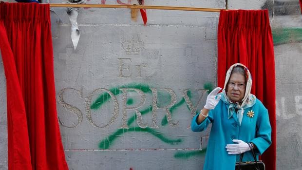 Un individuo disfrazado de la reina Isabel II gesticula con la obra de Banksy detrás
